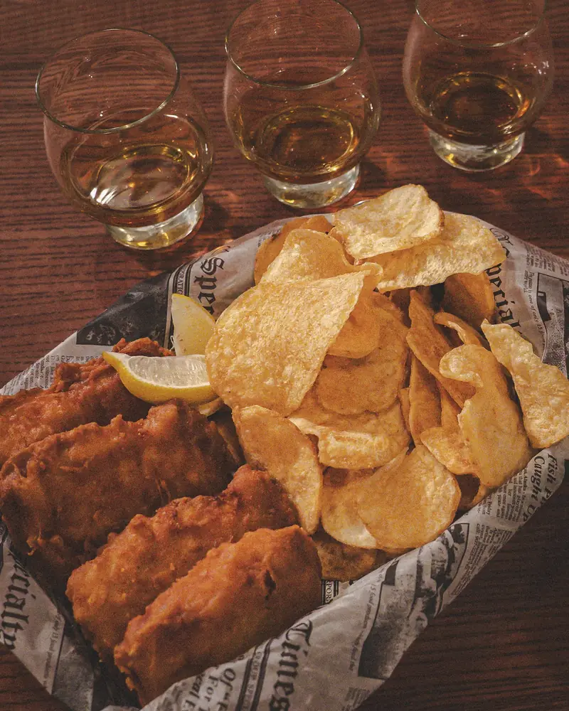 Fish and chips in a newspaper-lined basket, a whiskey flight behind it