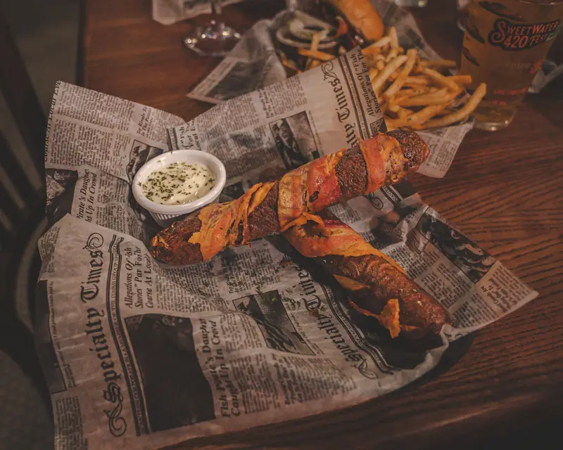 Two bacon-wrapped pretzel rods with a dip, in a newspaper-lined basket